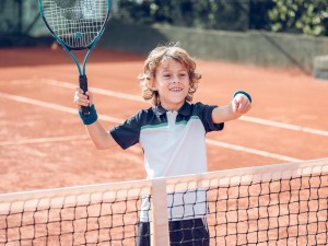 Cheerful little boy with curly blond hair in sportswear smiling brightly and pointing away while standing near net on court with racket during tennis training